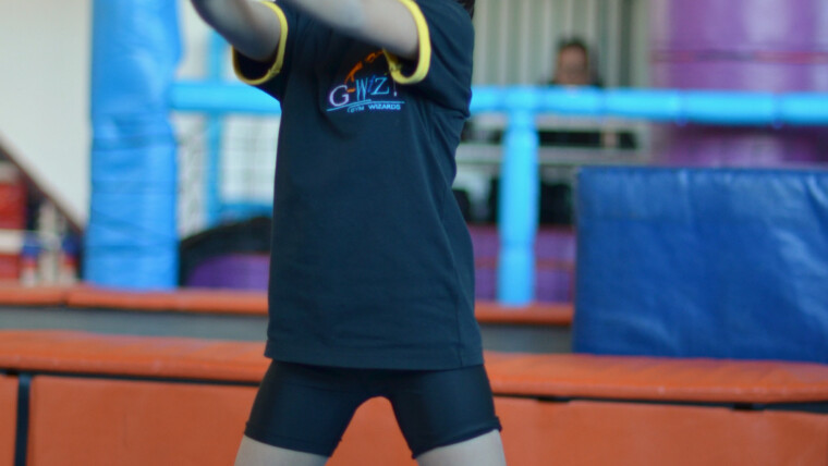 Girl in a Gym Wizards T-shirt standing with feet wide, reaching both hands to one side so one arm is crossing the midline during a warm-up